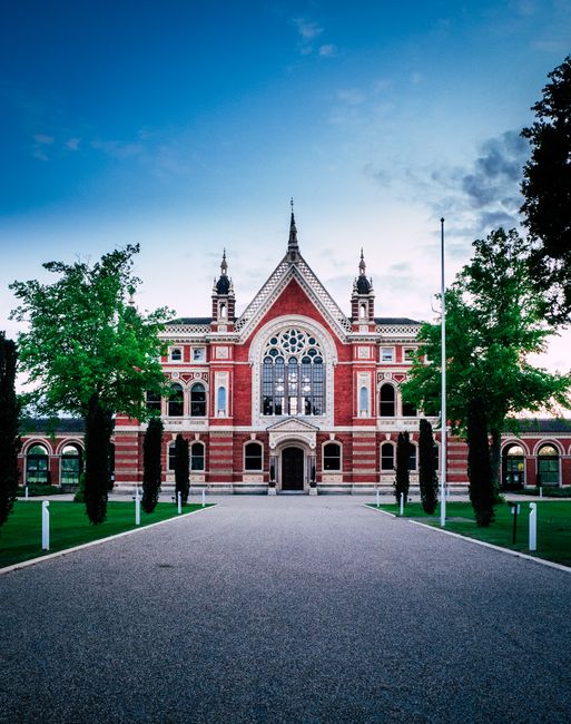 A historic red brick building with ornate architectural details, including decorative trim, large arched windows with stained glass, and spire-like towers on either side, situated behind a paved walkway lined with evenly spaced trees and white posts. The building is set against a partly cloudy sky with soft daylight, and there are no people or vehicles visible. The scene is captured as part of a house removal or furniture transport process near Dulwich Picture Gallery in Dulwich, with the setting suggesting preparation for a relocation or moving service by Man With a Van Dulwich, focused on logistics and packing for house removals.