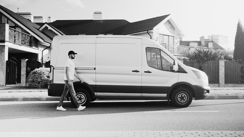 A man wearing a white t-shirt, dark pants, and a cap is walking alongside a large white cargo van parked on a residential street in East Dulwich. The van, which is part of the services offered by Man With a Van Dulwich, is positioned parallel to the sidewalk with its side door closed. In the background, there are modern residential houses with pitched roofs, large windows, and balconies, along with landscaped gardens and fences. The street surface is visible with painted road markings, and the scene suggests a home relocation or furniture transport process. The overall environment appears to be a quiet suburban area during daylight, with natural light illuminating the scene. The van is equipped for loading and moving household furniture, and the setting reflects professional removals and packing activities associated with house moves, as managed by [COMPANY_NAME].