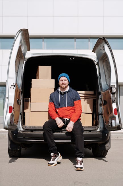 A young man with a beard, wearing a blue beanie, a red and navy blue jacket, black pants, and white sneakers, is sitting on the ground in front of an open van with its rear doors wide open. Inside the van, there are multiple neatly stacked cardboard boxes of various sizes, some wrapped in plastic for protection. The vehicle is positioned in an outdoor setting on a paved surface, with a modern building featuring large windows and white wall panels in the background. The scene captures the loading process for a home relocation, illustrating furniture transport and packing elements as part of a professional removals service. The presence of the boxes, the open van ready for loading or unloading, and the person's relaxed posture all reflect typical steps involved in furniture transport and moving logistics, consistent with services offered by Man With a Van Dulwich.