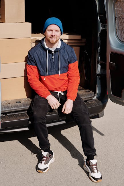 A man wearing a white t-shirt, dark pants, and a cap is walking alongside a large white cargo van parked on a residential street in East Dulwich. The van, which is part of the services offered by Man With a Van Dulwich, is positioned parallel to the sidewalk with its side door closed. In the background, there are modern residential houses with pitched roofs, large windows, and balconies, along with landscaped gardens and fences. The street surface is visible with painted road markings, and the scene suggests a home relocation or furniture transport process. The overall environment appears to be a quiet suburban area during daylight, with natural light illuminating the scene. The van is equipped for loading and moving household furniture, and the setting reflects professional removals and packing activities associated with house moves, as managed by [COMPANY_NAME].