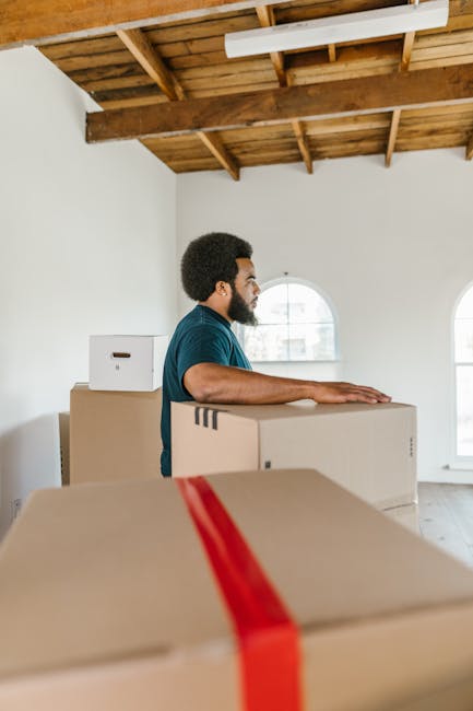 A man with dark curly hair and a beard, dressed in a teal t-shirt, is standing inside a spacious room with a wooden ceiling and two arched windows, preparing for a home relocation. He is positioned behind several cardboard boxes of varying sizes, which are stacked on top of each other. The boxes are taped and ready for transport, with one box featuring red packing tape. The man appears focused, with his hands resting on one of the boxes, indicating the process of packing or organizing for a move. The room has natural lighting coming through the windows, and the environment suggests a professional move or packing service, such as those offered by Man With a Van Dulwich, supporting furniture transport, packing, and loading processes in a typical house removal scenario.