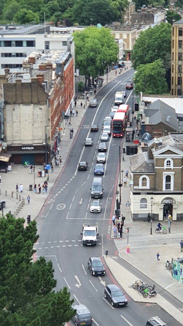 An aerial view of Lordship Lane in Dulwich showing a busy street with multiple vehicles, including cars, vans, and buses, moving along the road, which is lined with a mix of commercial and residential buildings. On the sidewalks, pedestrians are walking and some are crossing the street at designated crossings. There are trees providing greenery along the pavement and in front of shops, creating a pleasant environment for a home relocation or furniture transport process. The image captures the urban setting during daylight, with clear visibility of the street layout and surrounding architecture, illustrating the typical environment in which Man With a Van Dulwich provides removals and moving services, including packing, loading, and transporting furniture and household items typical of house removals in the area.
