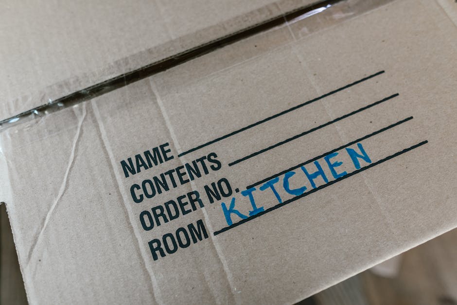 A man with dark curly hair and a beard, dressed in a teal t-shirt, is standing inside a spacious room with a wooden ceiling and two arched windows, preparing for a home relocation. He is positioned behind several cardboard boxes of varying sizes, which are stacked on top of each other. The boxes are taped and ready for transport, with one box featuring red packing tape. The man appears focused, with his hands resting on one of the boxes, indicating the process of packing or organizing for a move. The room has natural lighting coming through the windows, and the environment suggests a professional move or packing service, such as those offered by Man With a Van Dulwich, supporting furniture transport, packing, and loading processes in a typical house removal scenario.