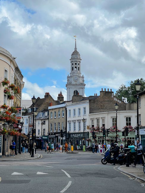 A busy street scene in Dulwich Village, with a historic white clock tower with a spire visible in the background under a partly cloudy sky. The foreground shows a pavement with pedestrians walking and standing near shopfronts, some engaging in household shopping or errands. Several storefronts with signage and window displays line the street, including flower baskets hanging from lampposts. A motorcycle and a small vehicle are parked on the streetside, while a van is partially visible on the right, indicating a moving or loading process. Behind, residential buildings with varied architectural styles and chimneys are visible, situating the scene within a typical UK residential and commercial area. This setting reflects the environment where household items may be packed, loaded, and transported during house removals or relocation services, with some equipment like shopping bags and packaging materials subtly present as part of the moving activity facilitated by Man With a Van Dulwich.