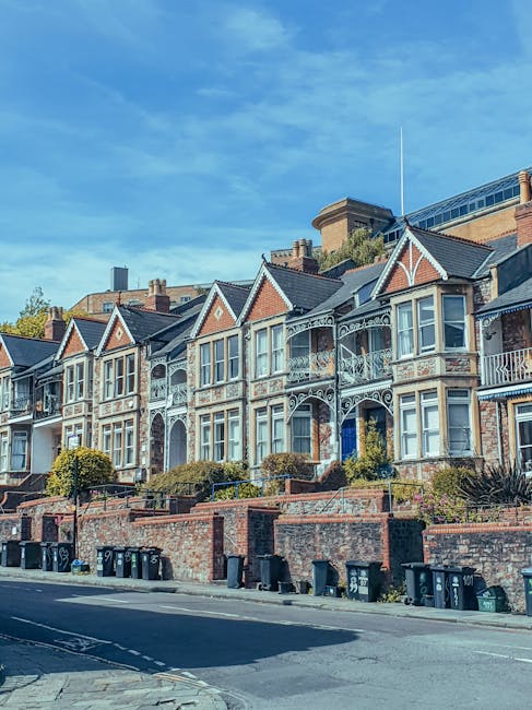 A row of Victorian-style terraced houses in Dulwich Village, featuring ornate white metalwork on the balconies and decorative window frames. The houses are built with red brick and have pitched roofs with chimneys, set behind a low brick wall with small landscaped gardens and bushes in front. The sidewalk in front is lined with black wheelie bins, and the street is quiet with no people visible. The sky above is blue with some wispy clouds, and the scene highlights a residential area suitable for home relocation or moving services in Dulwich, as offered by Man With a Van Dulwich.
